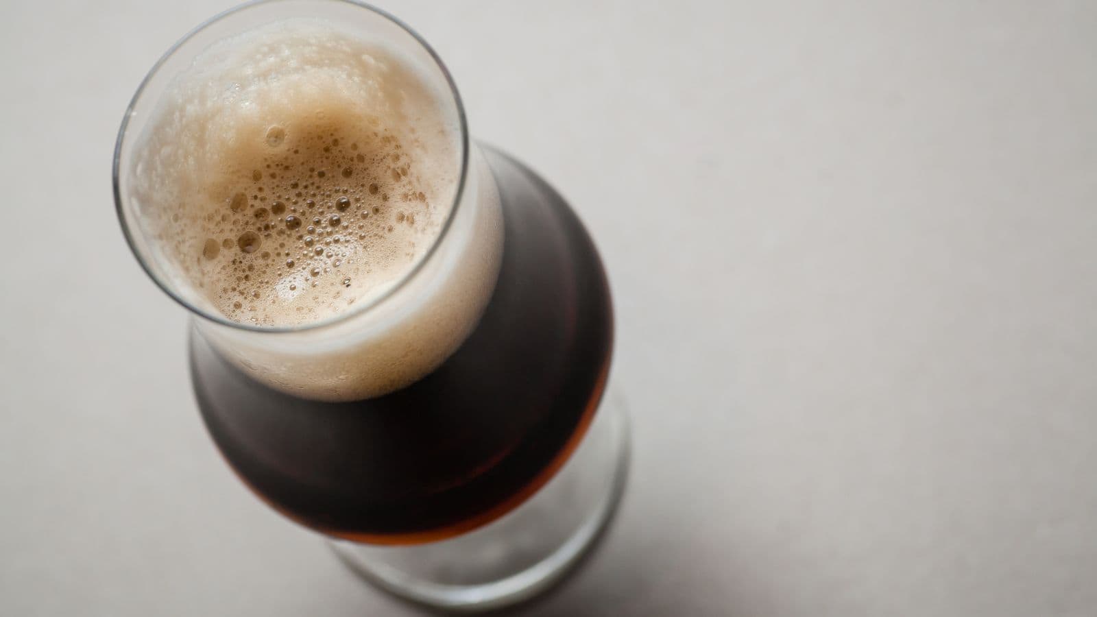 A close-up image of a dark beer in a glass on a light gray surface. The top of the beer is covered with a layer of frothy foam. The glass is viewed from above, highlighting the contrast between the beer's dark color and the creamy foam.