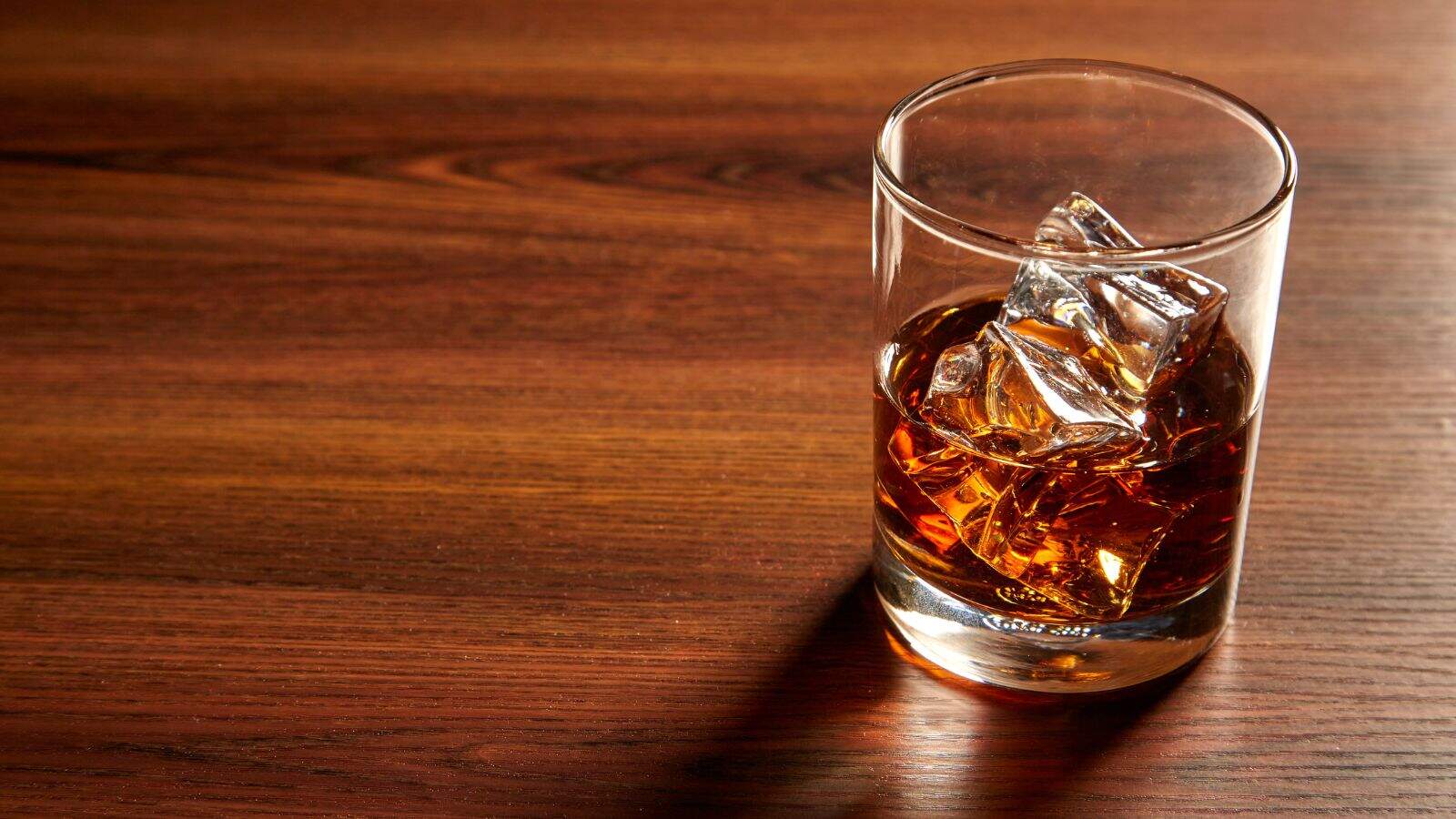 A glass of whiskey with ice cubes sits on a wooden surface, casting a shadow to the left. The drink has an amber color, and the ice is partially melted.