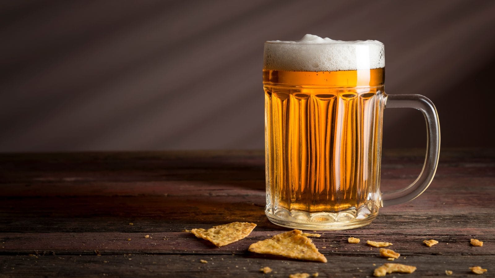 A glass mug filled with frothy amber beer sits on a wooden table. Crumbs and pieces of cracker are scattered around the base of the mug. The background is dimly lit with soft, diagonal shadows.