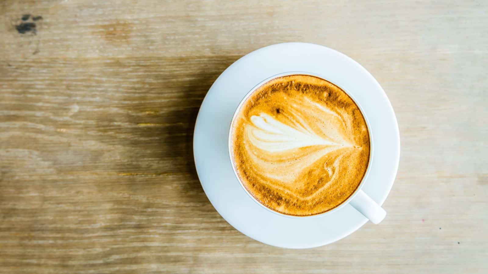 A white cup filled with a cappuccino, featuring a heart-shaped latte art design on top. The cup is placed on a matching saucer, and the background is a light wooden surface.