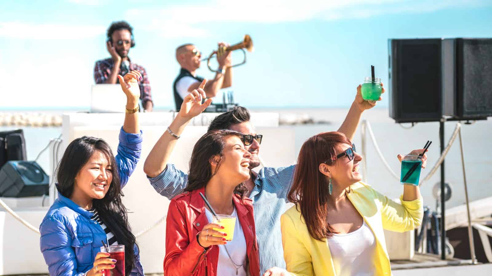 A group of four people holding drinks are dancing and smiling at an outdoor event near the water. Behind them, a DJ and a trumpet player perform on a stage. It is sunny, and everyone appears to be enjoying the music.