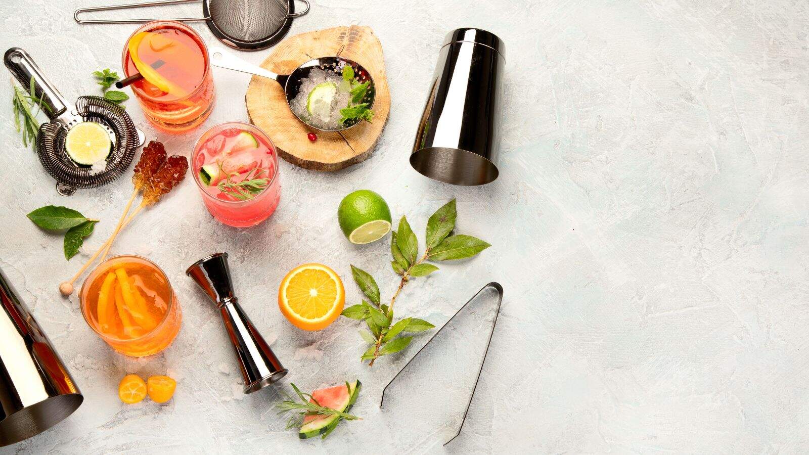 Various cocktail-making tools, three colorful drinks with garnishes, sliced citrus fruits, ice, mint leaves, and a jigger are arranged on a light textured surface with space on the right.