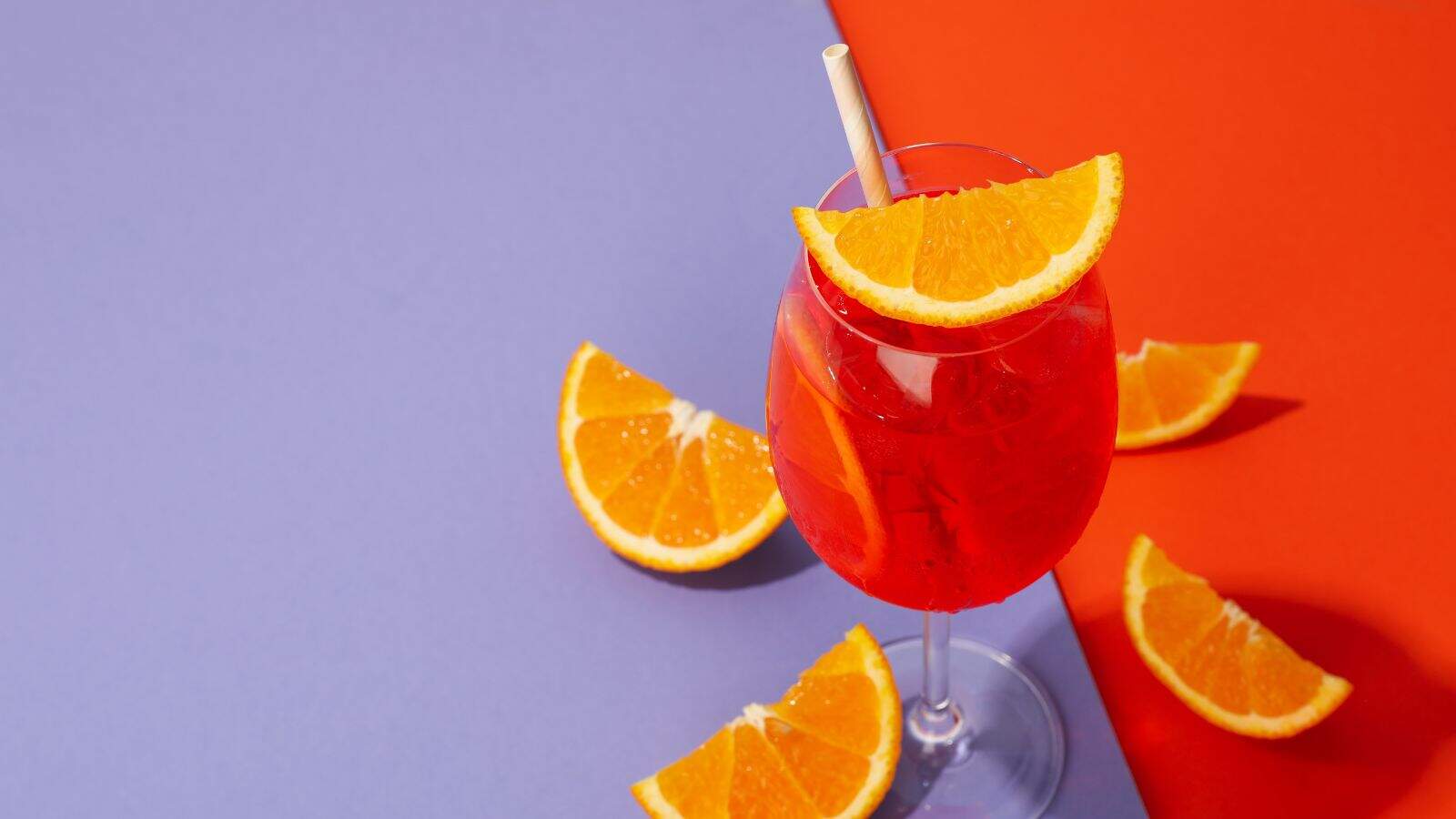 A glass filled with a red-orange beverage, garnished with a slice of orange and a straw, sits on a surface divided into purple and red areas, with several additional orange slices scattered around.