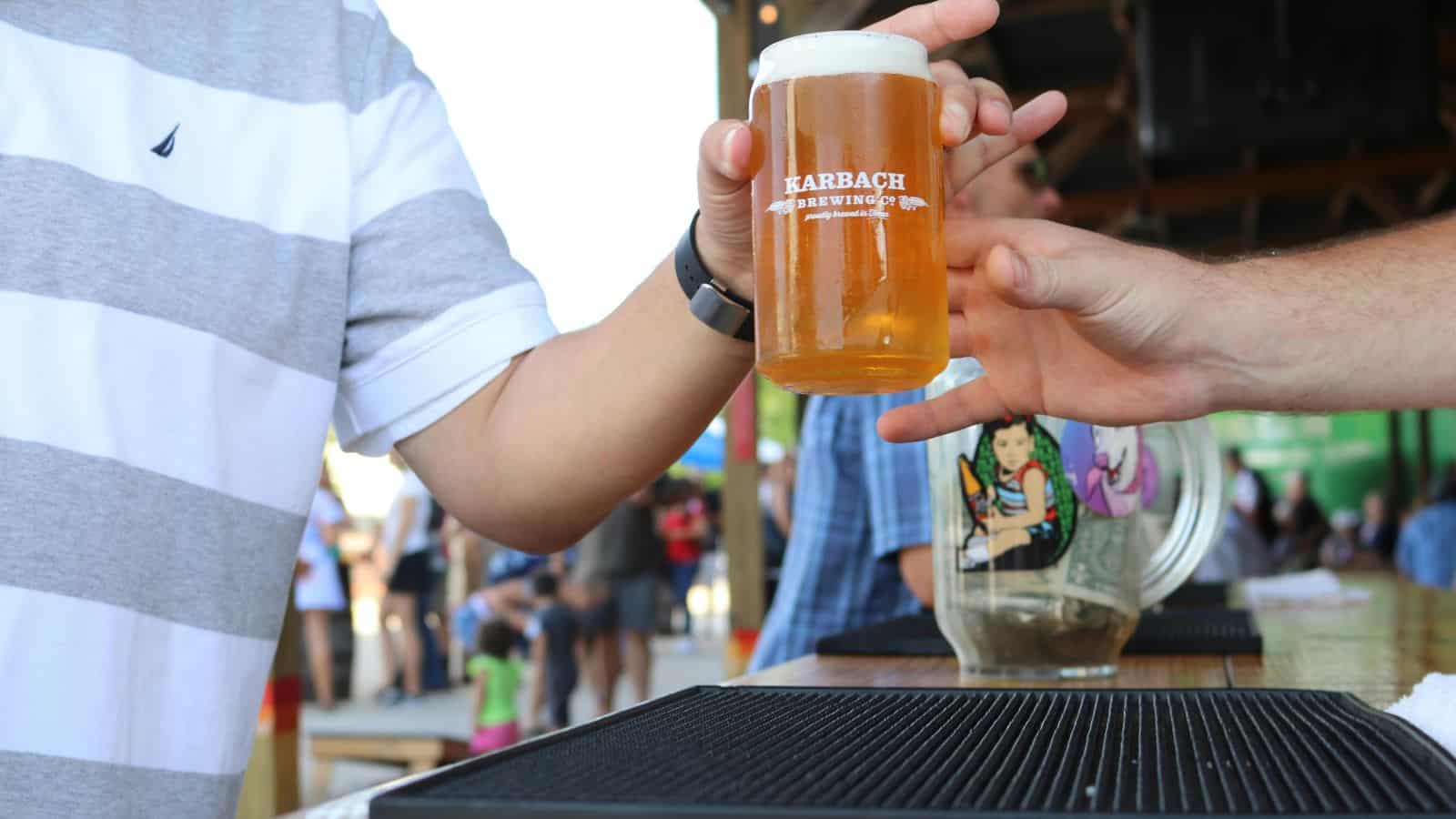 A person in a striped shirt receives a pint glass of beer from another person at an outdoor bar, with people and a colorful mural visible in the blurred background.