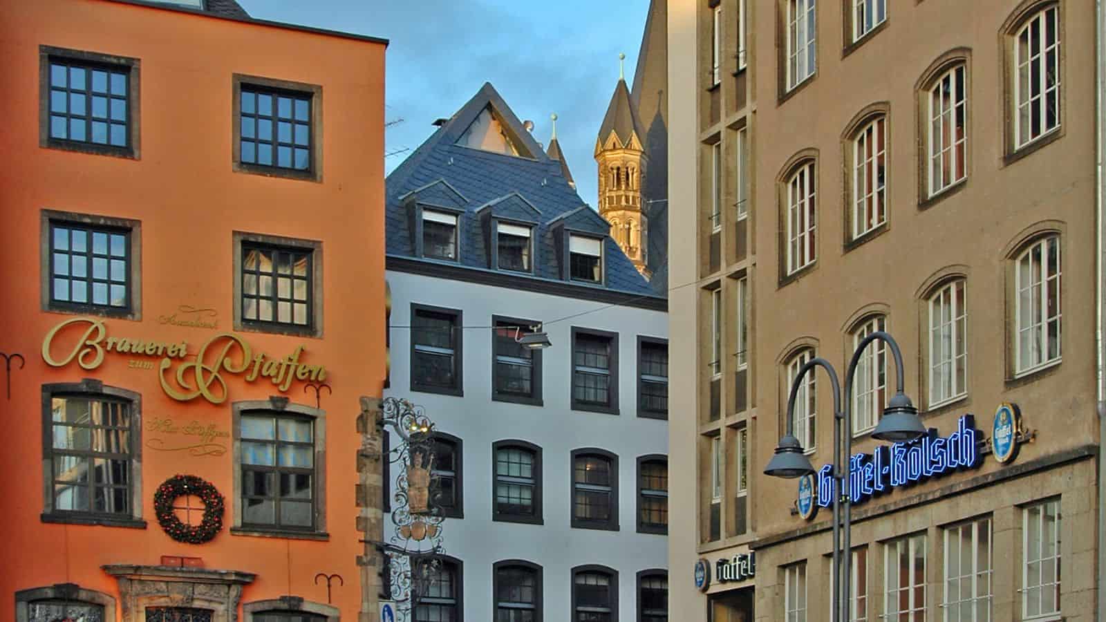 Street view of colorful buildings in a European city. An orange building features signs reading "Brauer" and "Cafe-Pub" with wreath decoration. A beige building has a blue sign. A cathedral spire is visible in the background under a cloudy sky.