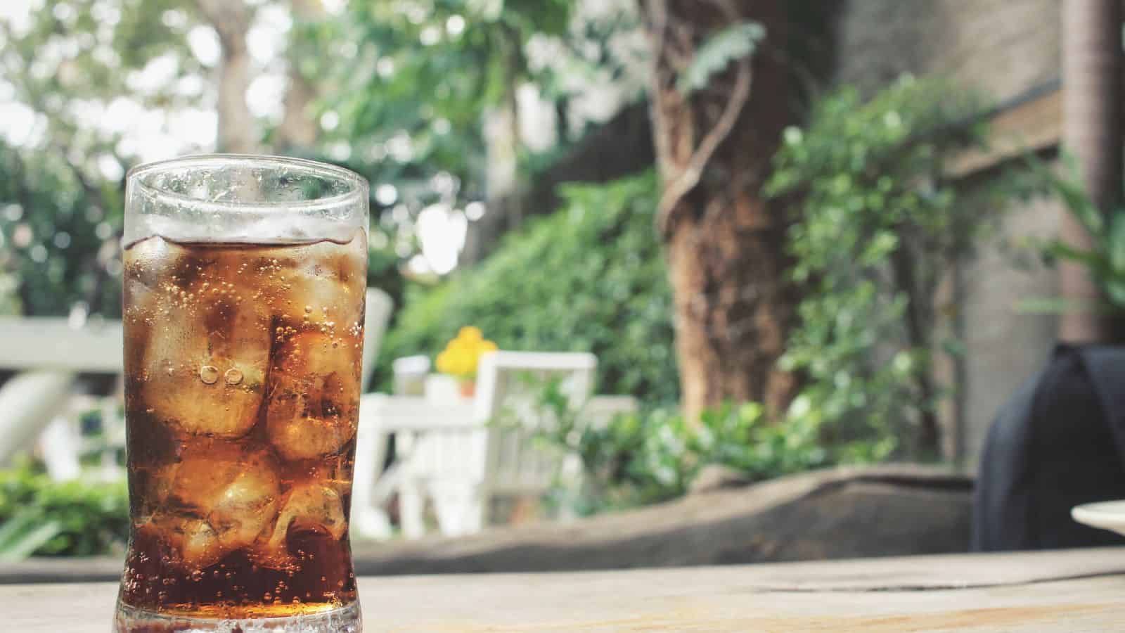 A glass filled with cola and ice cubes sits on a wooden table. The background features blurred greenery and patio furniture, suggesting an outdoor setting.