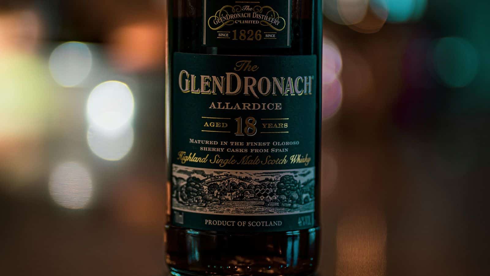 A close-up of a bottle of GlenDronach Allardice 18-year-old Highland single malt Scotch whisky, showing the front label with product details and a blurred background of lights.