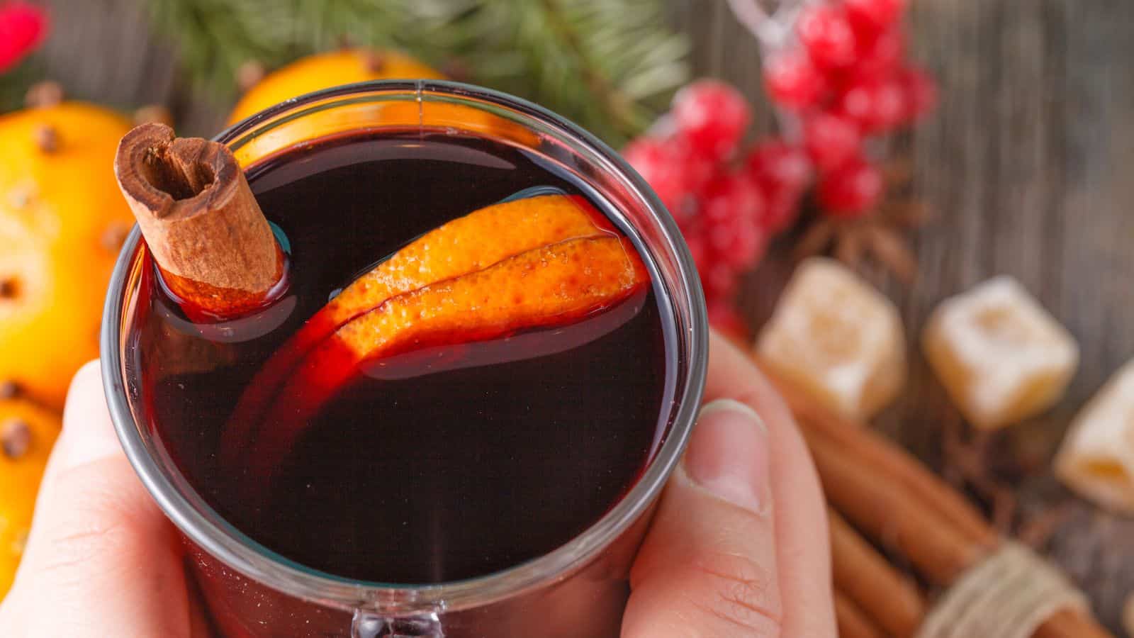 A person holds a glass mug of mulled wine garnished with a cinnamon stick and orange slices. The background features red berries, orange slices, cinnamon sticks, and sugar cubes on a wooden surface.