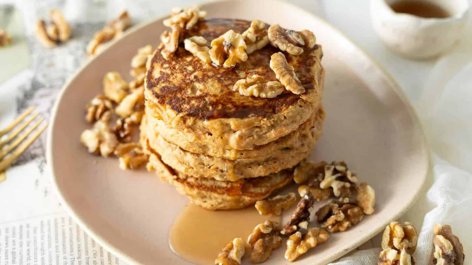 A stack of three pancakes topped with walnuts is on a pink plate. Syrup is being drizzled over the pancakes. Additional walnuts are scattered around the plate. Two carrots are partially visible in the background.