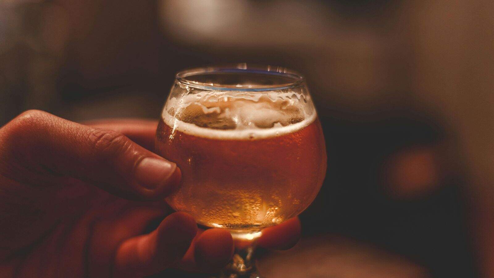 A hand holds a small, round glass filled with amber-colored beer. The glass has a frothy head on top. The background is softly blurred, emphasizing the beverage.
