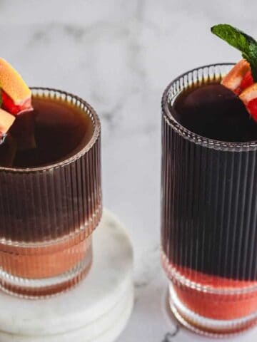 Two ribbed glasses filled with a dark beverage, each garnished with a slice of grapefruit and a sprig of mint, are placed on a light marble surface, with one glass set on stacked marble coasters.