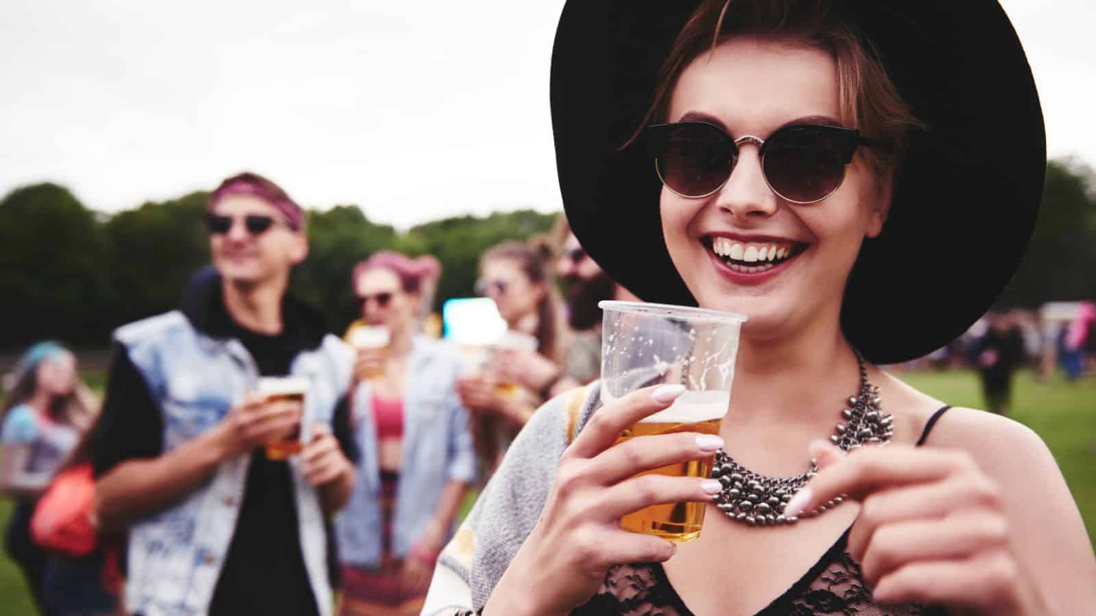 A person wearing sunglasses and a black hat smiles while holding a plastic cup with a drink. Several people in casual clothing are gathered in the background at an outdoor event.