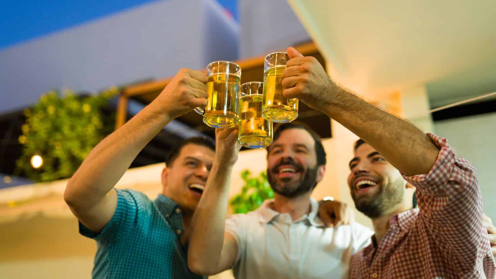 Three men are smiling and holding up glasses of beer in a toast, standing together outdoors under a covered patio area with trees and buildings in the background.