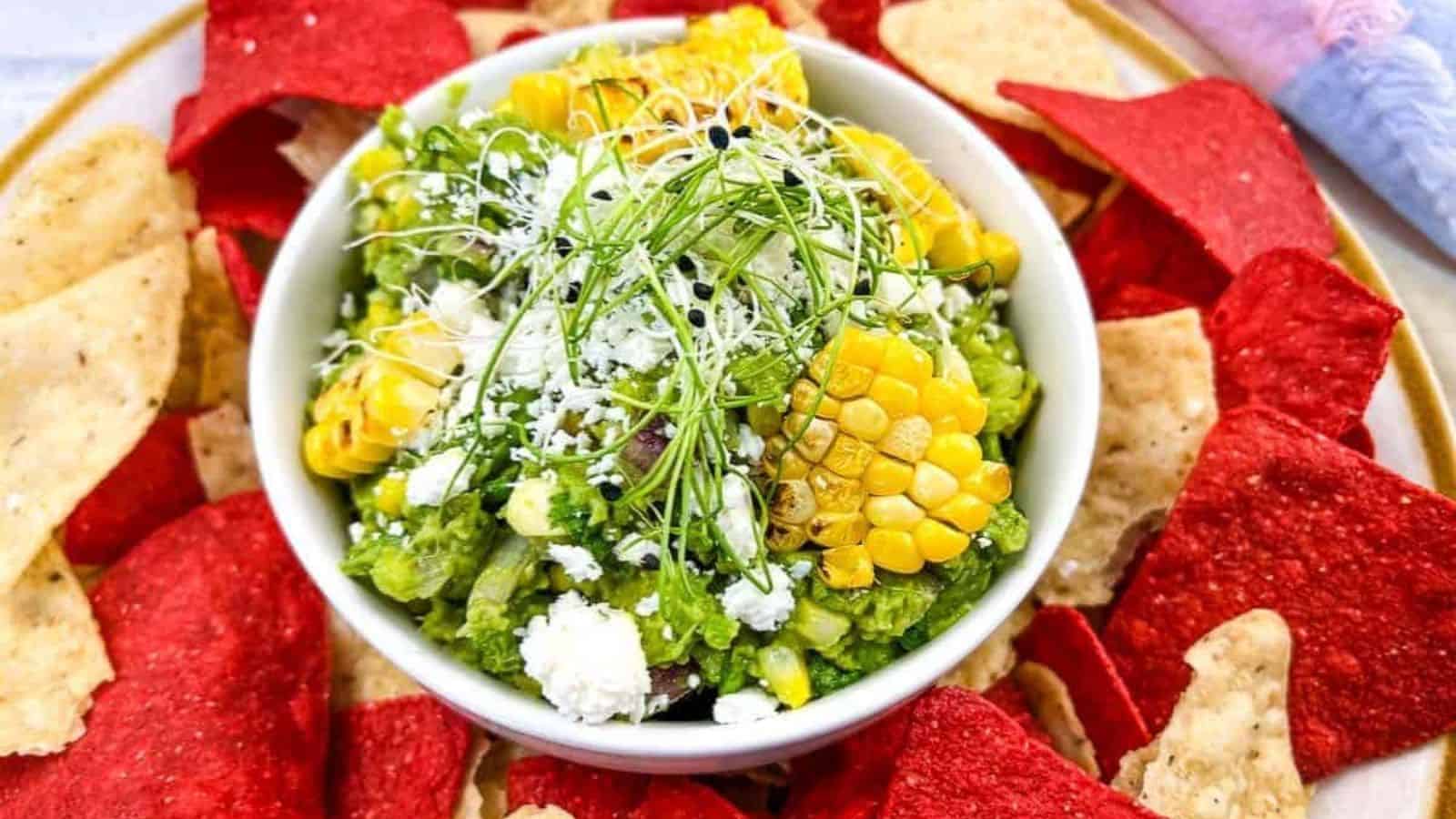A bowl of guacamole topped with grilled corn, cheese, and sprouts, surrounded by red and yellow tortilla chips on a plate.