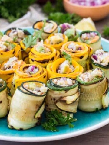 A blue plate with neatly arranged zucchini and yellow squash roll-ups filled with diced vegetables and herbs. Small bowls of ingredients and fresh parsley are visible in the background.