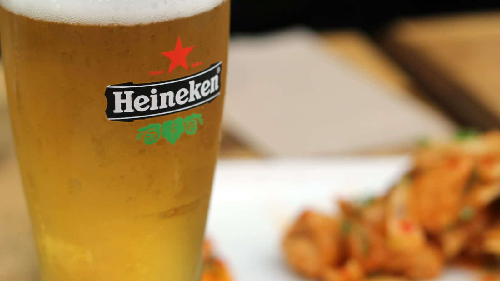 A close-up of a glass of Heineken beer with condensation on the outside, set on a table next to a plate of food in the background.