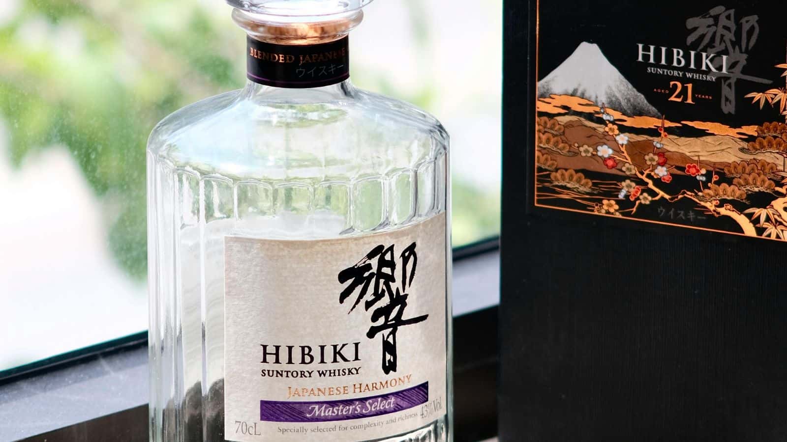 A close-up of a Hibiki Suntory Whisky bottle labeled "Japanese Harmony Master's Select" beside a black box with gold and red accents, including a mountain and floral design. Green foliage is blurred in the background.