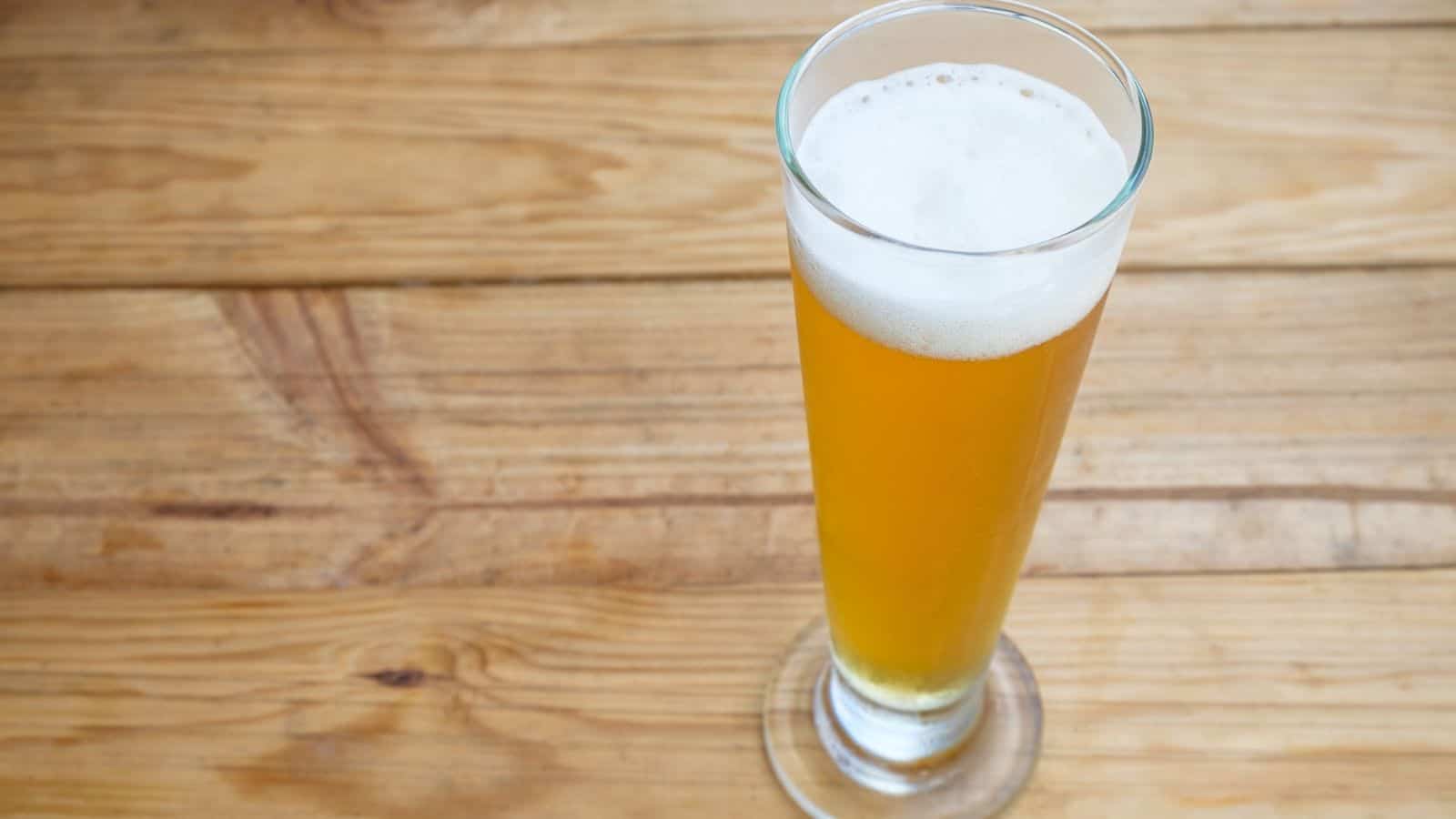 A tall glass of light amber beer with a frothy head sits on a wooden table.