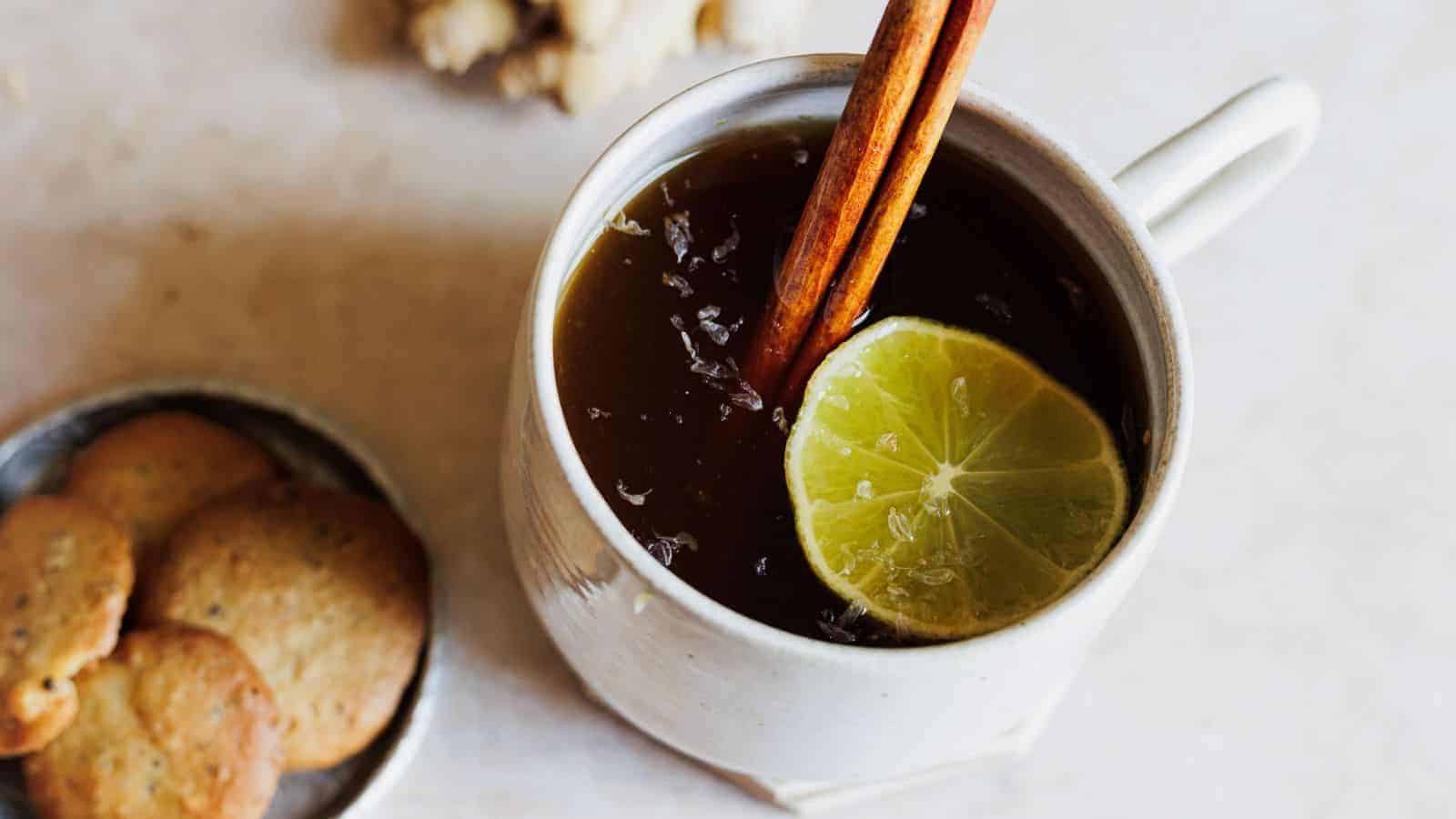 A mug filled with a dark beverage, garnished with a lime slice and a cinnamon stick, sits on a table. Next to it, a small plate holds several round cookies. A piece of ginger is visible in the background.