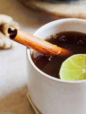 A white ceramic mug filled with a dark beverage, garnished with a cinnamon stick and a lime slice. A small piece of ginger is beside the mug, placed on a light-colored surface.