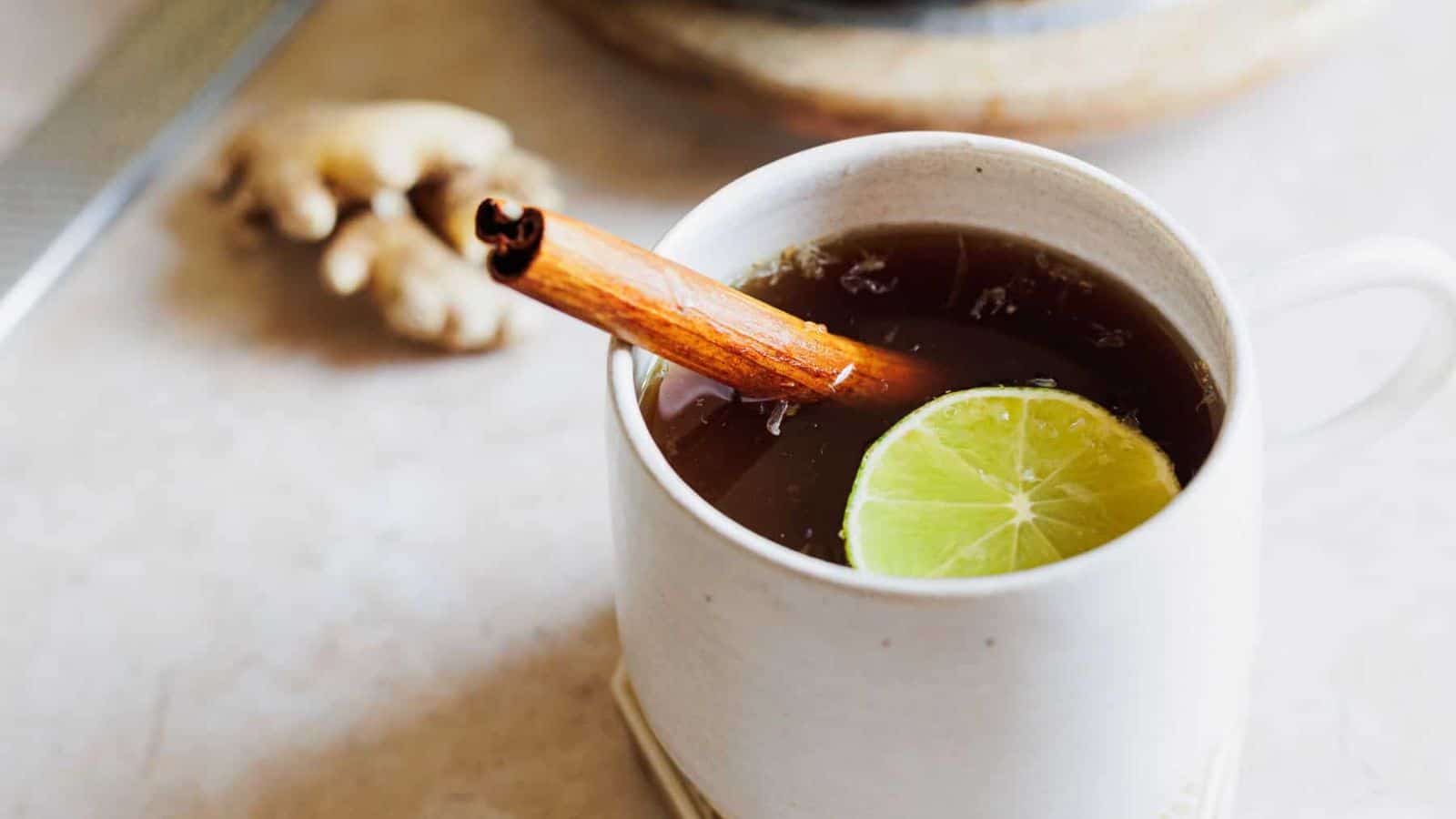 A white ceramic mug filled with a dark beverage, garnished with a cinnamon stick and a lime slice. A small piece of ginger is beside the mug, placed on a light-colored surface.