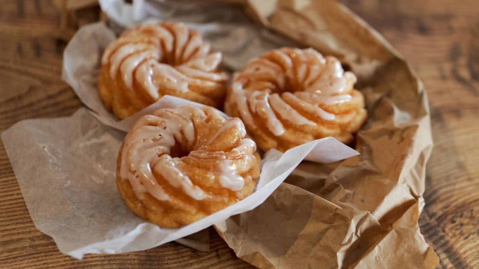 Three glazed bundt cakes rest on crumpled parchment paper atop a textured wooden surface. Each cake has a spiral design with a light glaze coating. The parchment paper wraps around the cakes, suggesting a freshly baked presentation.