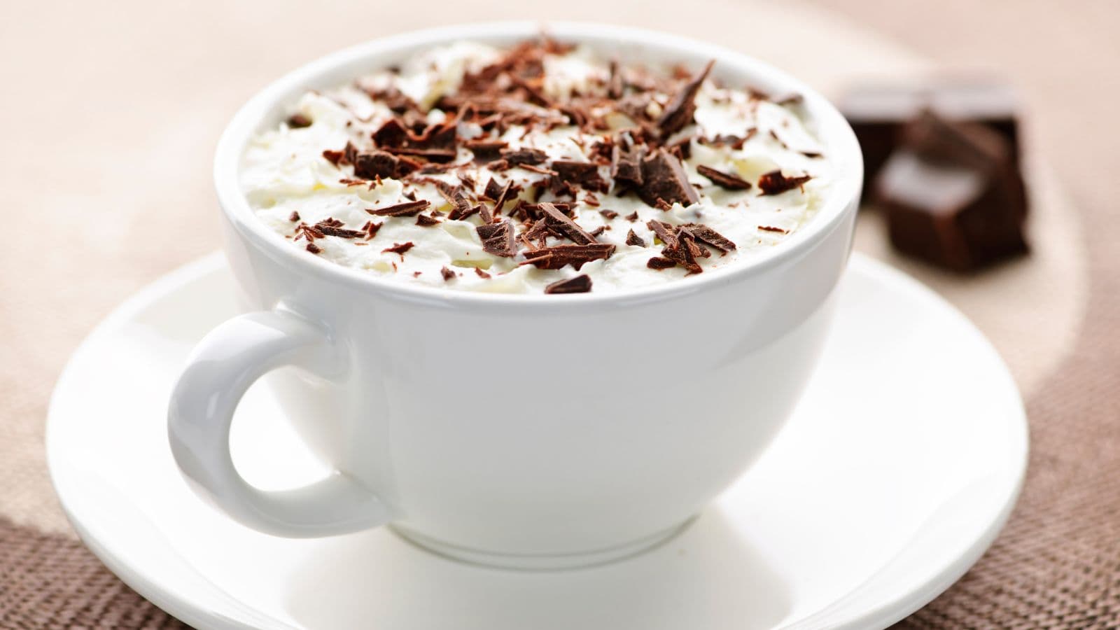 A white cup filled with a creamy beverage topped with whipped cream and chocolate shavings sits on a matching white saucer. Two pieces of chocolate are blurred in the background. The surface underneath is a textured brown material.