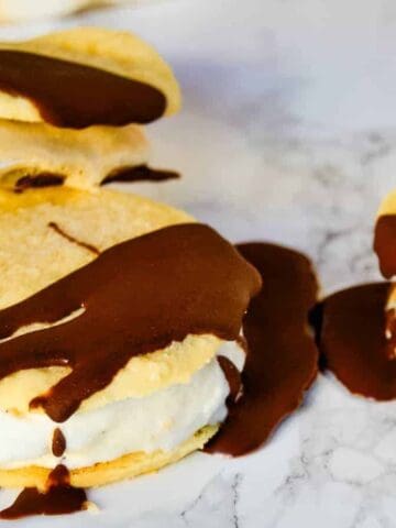 Three ice cream cookie sandwiches rest on a marble surface. Each sandwich consists of two cookies with ice cream in the middle, topped with hardened chocolate sauce drizzled over the top.