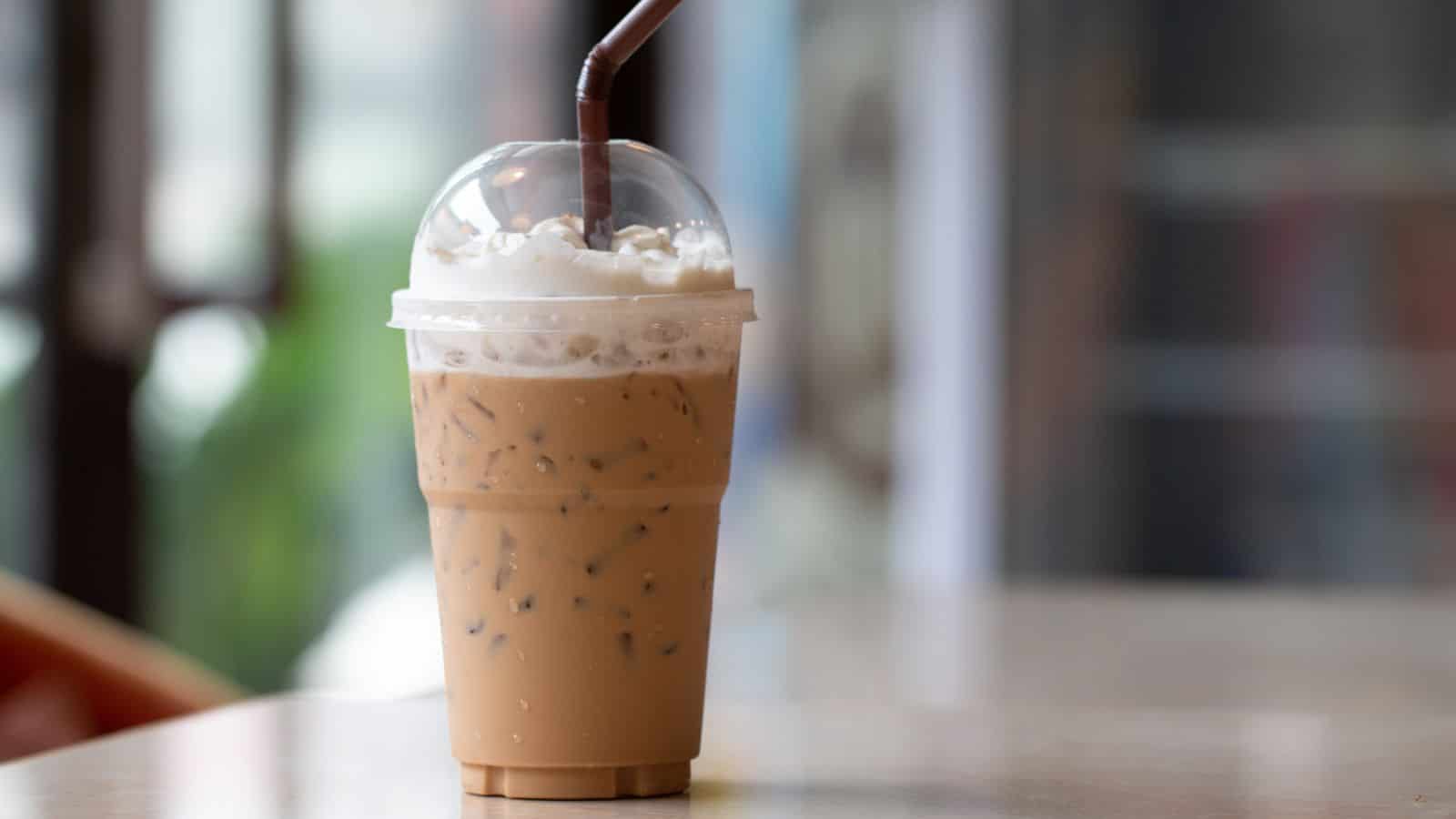 A clear plastic cup filled with iced coffee sits on a table. The drink has a light brown color, topped with a dome lid and whipped cream. A brown straw protrudes from the lid. The background is slightly blurred, suggesting an indoor setting.
