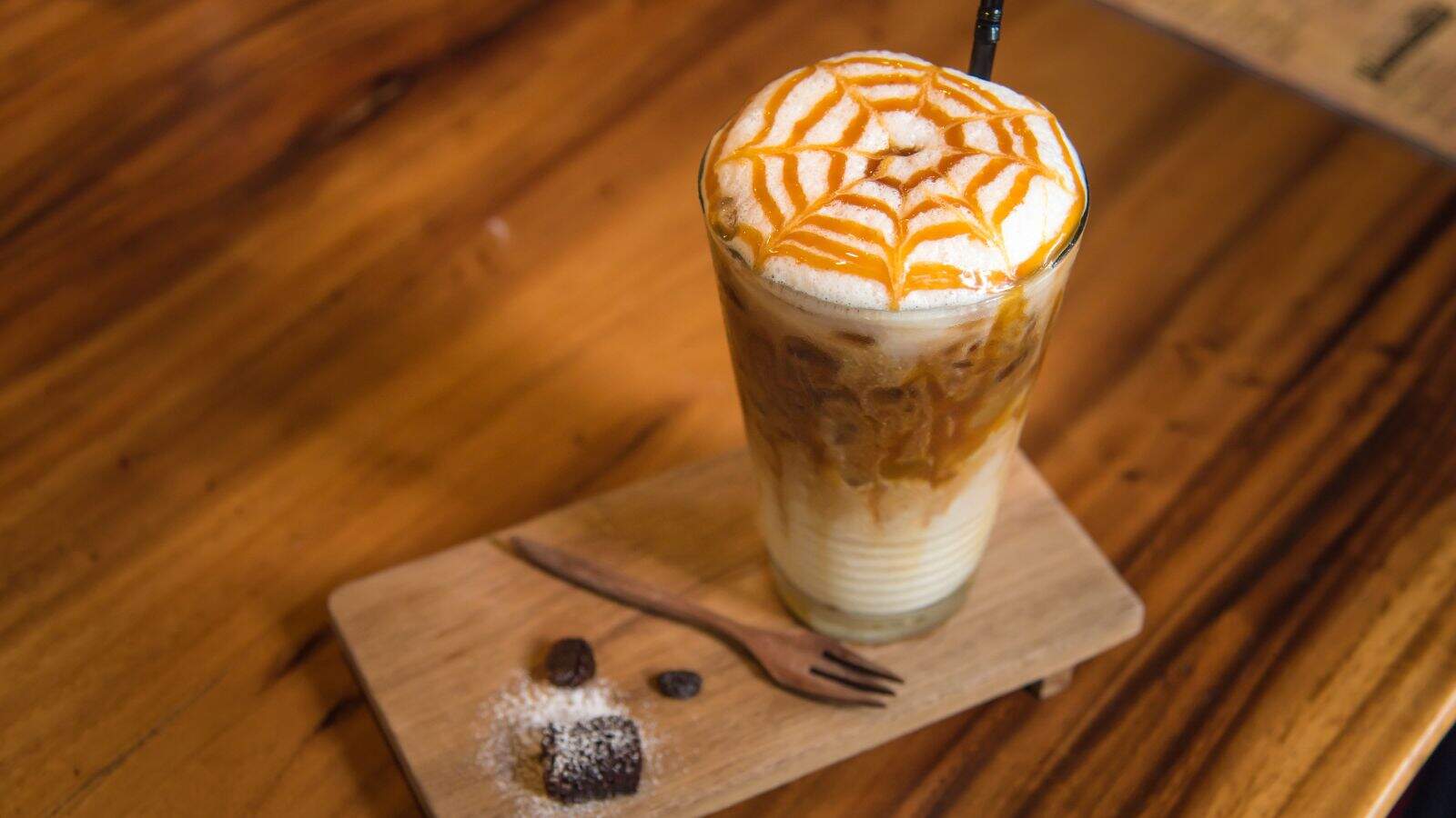 A tall glass of iced coffee sits on a wooden board. The drink features creamy layers topped with foamy milk and caramel drizzle in a spiderweb pattern. A reusable straw is in the glass. A small wooden fork and coffee beans are on the board.