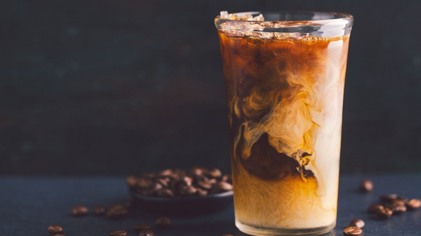A tall glass filled with iced coffee shows a swirling mix of dark coffee and light cream. Ice cubes float at the top. Coffee beans are scattered around a small bowl in the background on a dark surface.