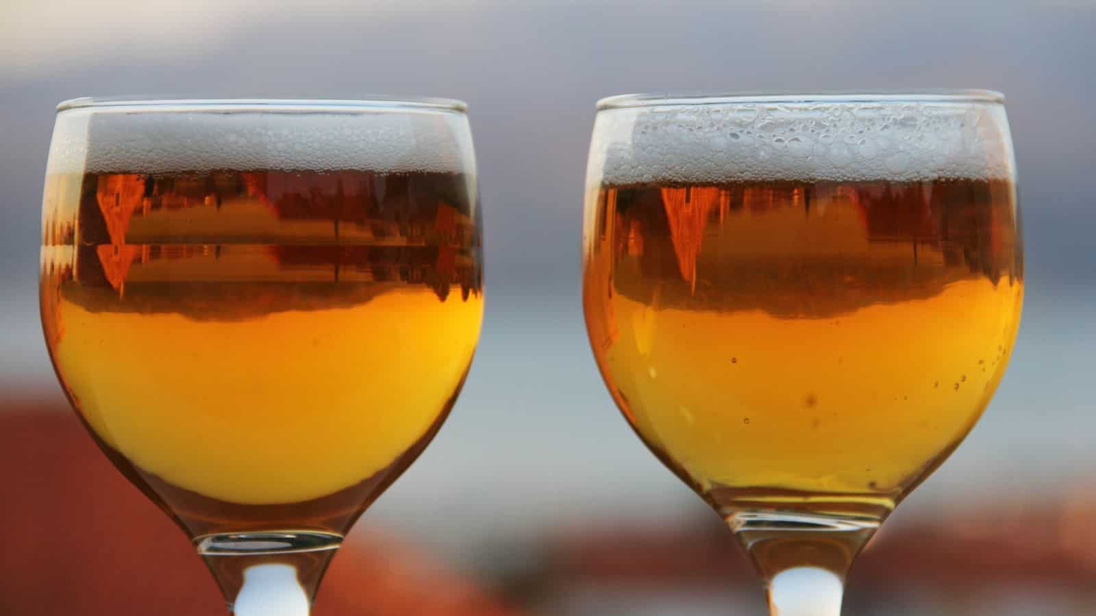 Two glasses filled with amber-colored beer are shown close up, with reflections visible on the glass surfaces. The background is blurred, showing a soft mix of colors.