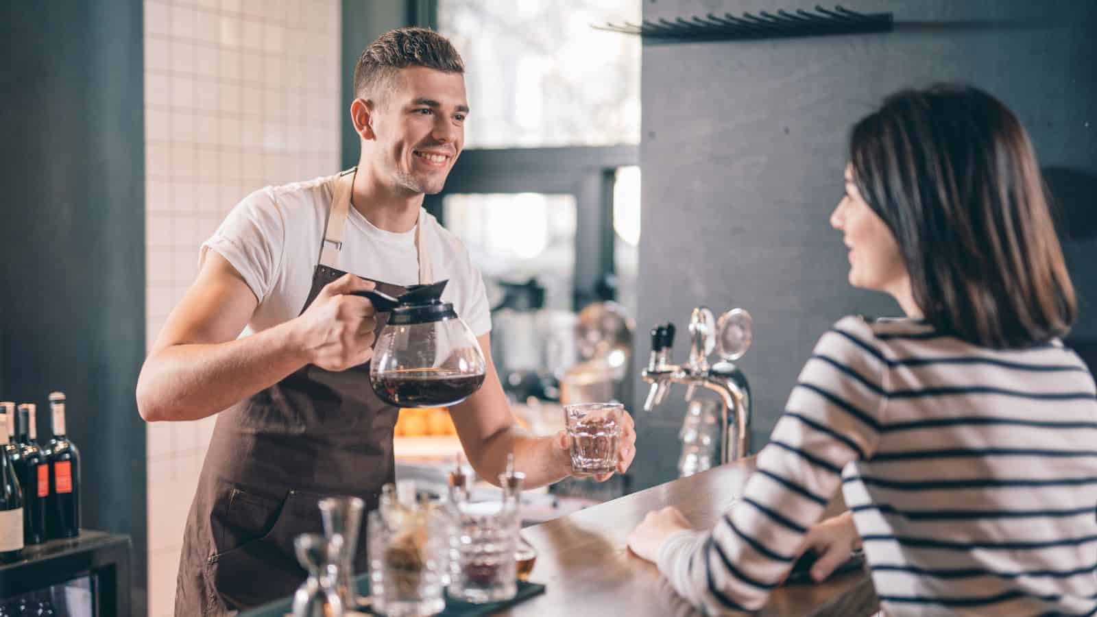 A barista in a white t-shirt and apron pours coffee into a glass mug for a customer at a cafe counter. The customer, wearing a striped shirt, smiles as she watches. Bottles and coffee-making tools are visible on the counter.