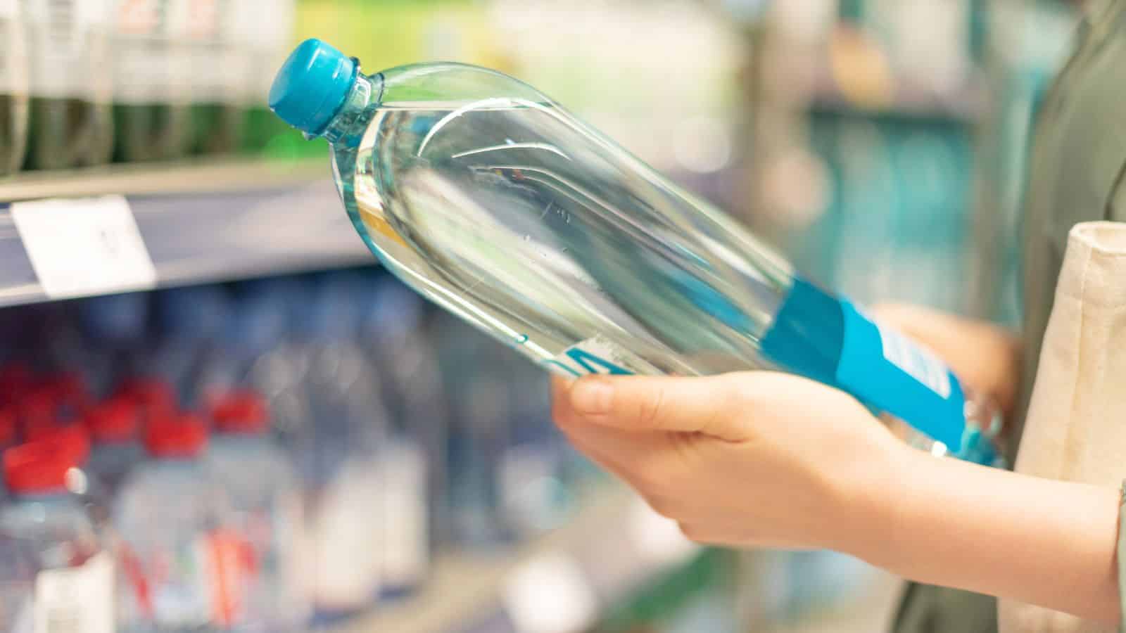 A person is holding a clear plastic bottle of water in a store aisle. The left hand supports the base, and the right hand holds the neck. There are shelves with various bottled items visible in the blurred background.