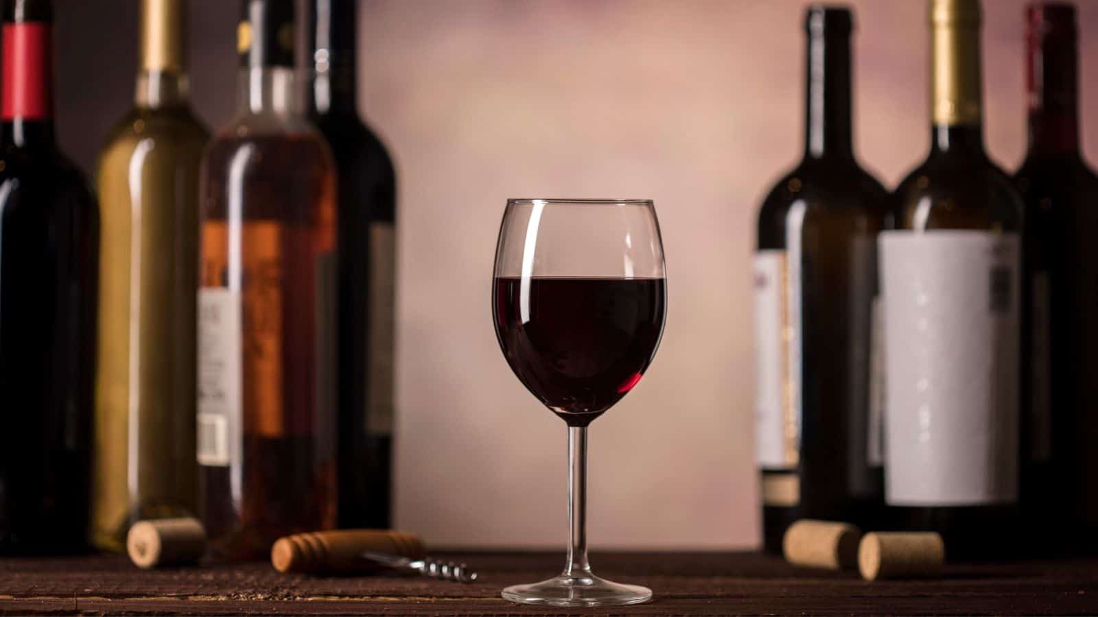 A glass of red wine is centered in front of several wine bottles, some opened with corks and a corkscrew on the wooden surface. The background is softly blurred, drawing focus to the wine glass.
