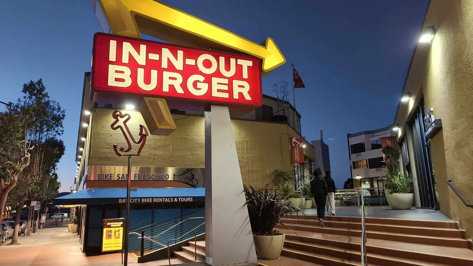 A large illuminated IN-N-OUT BURGER sign with a yellow arrow stands near stairs leading to the restaurant entrance at dusk; nearby are signs for bike rentals and tours.