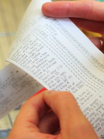 A person holds and examines a long grocery receipt with both hands. The printed text includes item prices and totals. Part of a shopping cart is visible in the background.