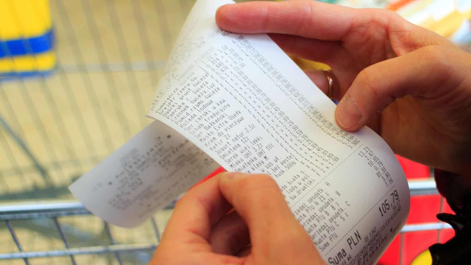 A person holds and examines a long grocery receipt with both hands. The printed text includes item prices and totals. Part of a shopping cart is visible in the background.