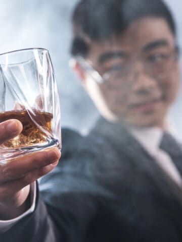 A man in a suit holds a glass of amber-colored liquor toward the camera with a blurred, smoky background. He is wearing glasses and appears to be holding a cigar in his other hand.