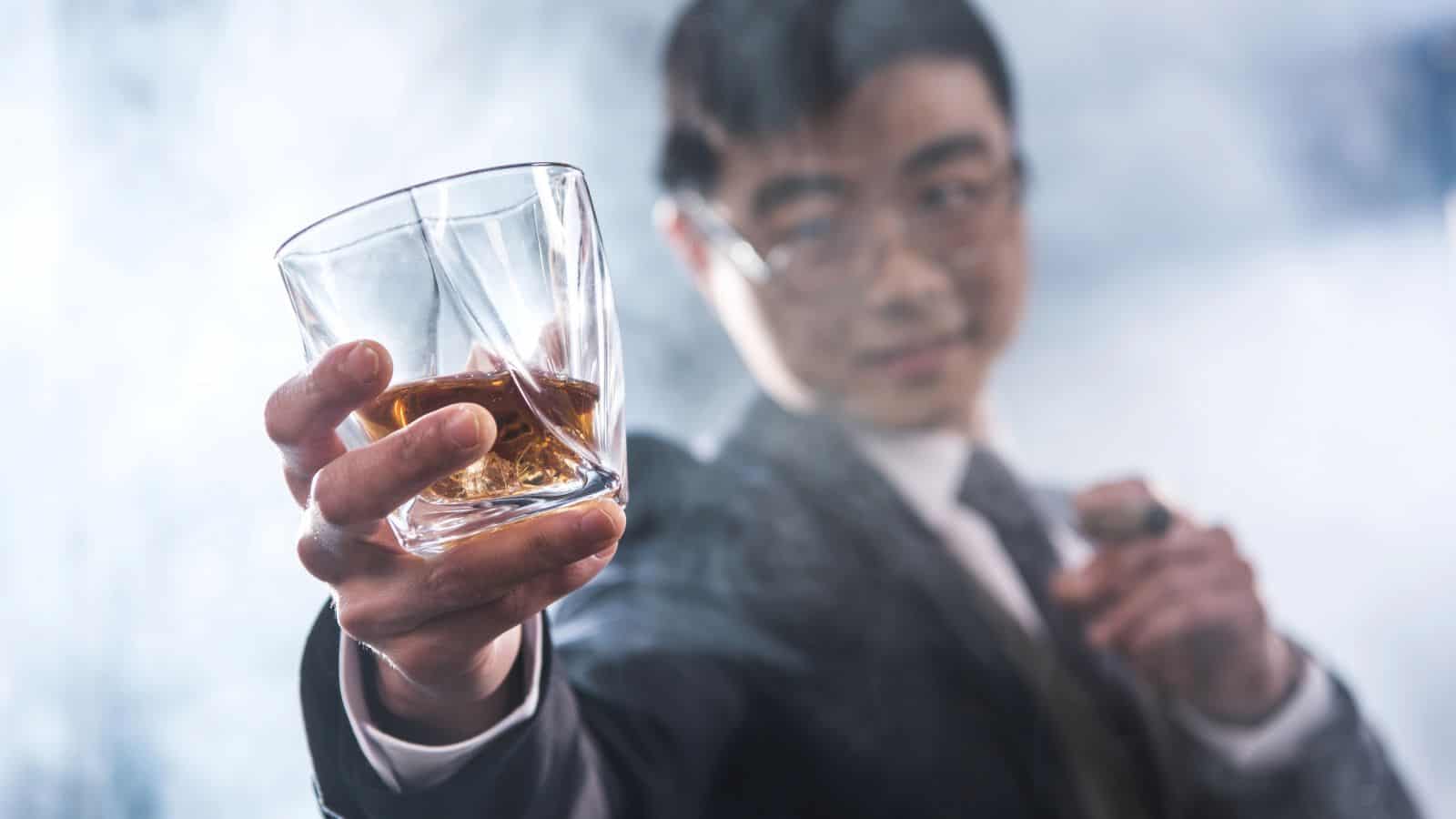A man in a suit holds a glass of amber-colored liquor toward the camera with a blurred, smoky background. He is wearing glasses and appears to be holding a cigar in his other hand.