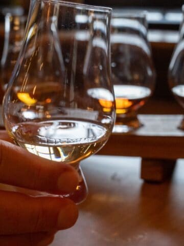 A hand holds a glass of whisky in the foreground, with a wooden tray holding three more whisky glasses in the background, all set on a wooden table.