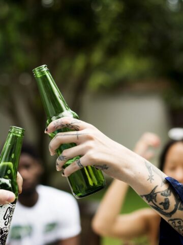 Two people are clinking green glass bottles together outdoors. Both have visible tattoos on their hands and arms. Others are blurred in the background, suggesting a casual outdoor gathering. Trees and greenery are visible.