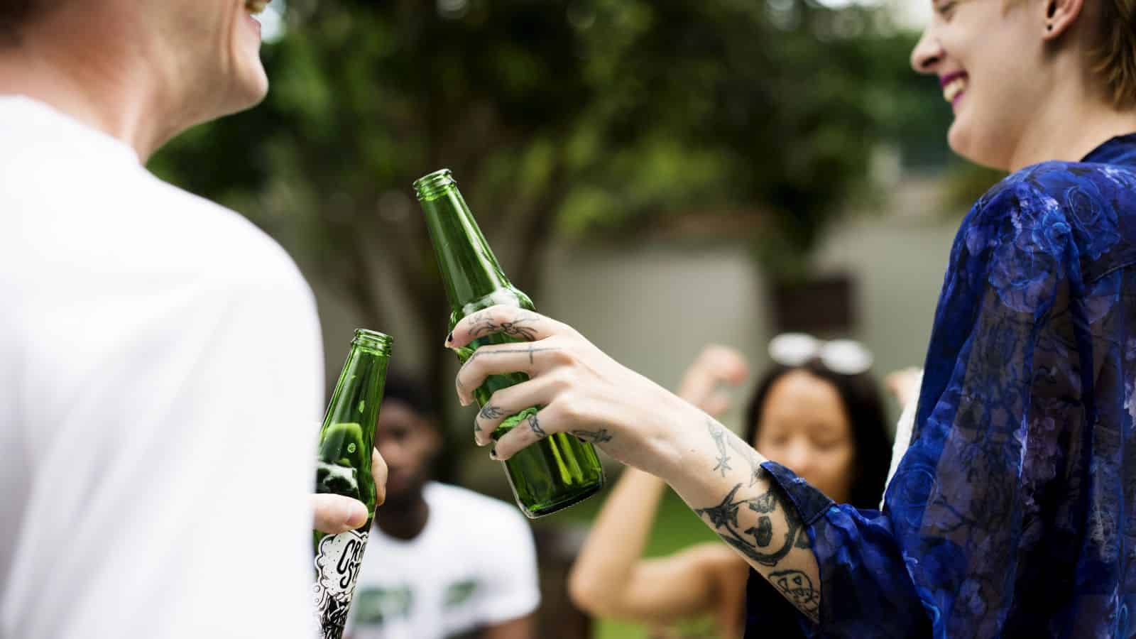 Two people are clinking green glass bottles together outdoors. Both have visible tattoos on their hands and arms. Others are blurred in the background, suggesting a casual outdoor gathering. Trees and greenery are visible.