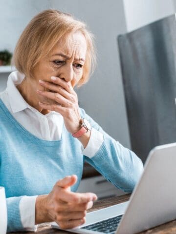 An older woman with short blond hair sits at a table in front of a laptop, covering her mouth with one hand and looking concerned. A white mug sits on the table beside her in a bright kitchen.
