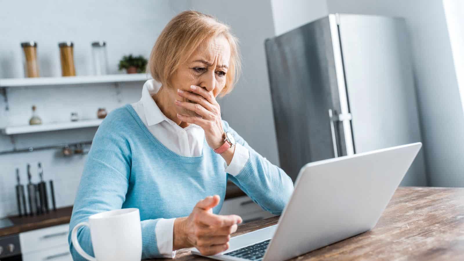 An older woman with short blond hair sits at a table in front of a laptop, covering her mouth with one hand and looking concerned. A white mug sits on the table beside her in a bright kitchen.
