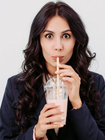 A woman with long dark hair in a dark blazer drinks a pink beverage through a straw from a plastic cup with a lid, sitting against a plain white background and looking directly at the camera with wide eyes.