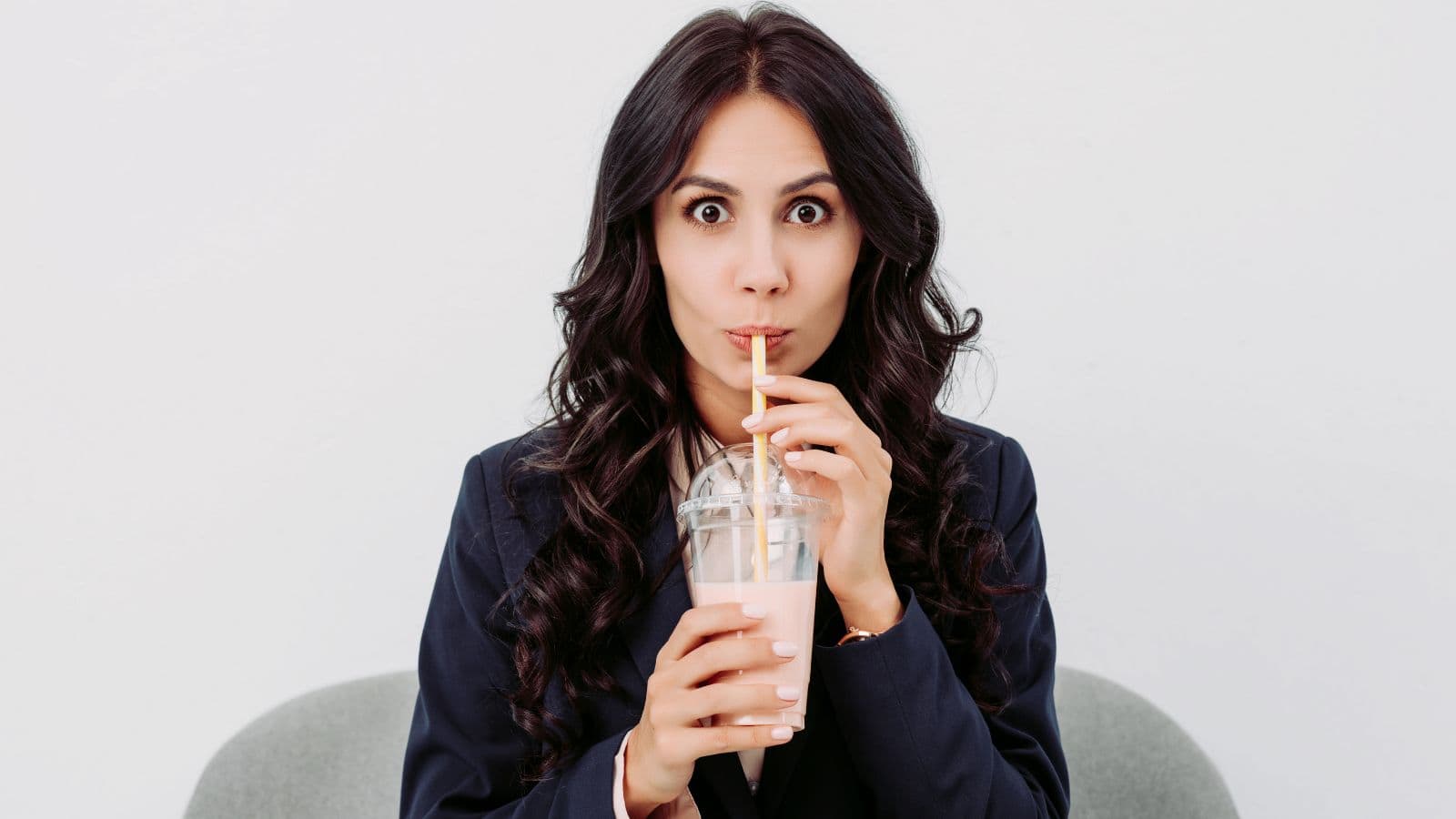 A woman with long dark hair in a dark blazer drinks a pink beverage through a straw from a plastic cup with a lid, sitting against a plain white background and looking directly at the camera with wide eyes.