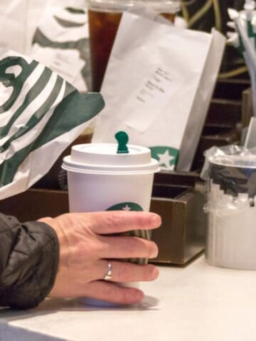 A person in a black jacket holds a Starbucks paper bag and a Starbucks cup at a counter. The counter also has a container of straws, a ceramic mug, and a plastic-wrapped item.