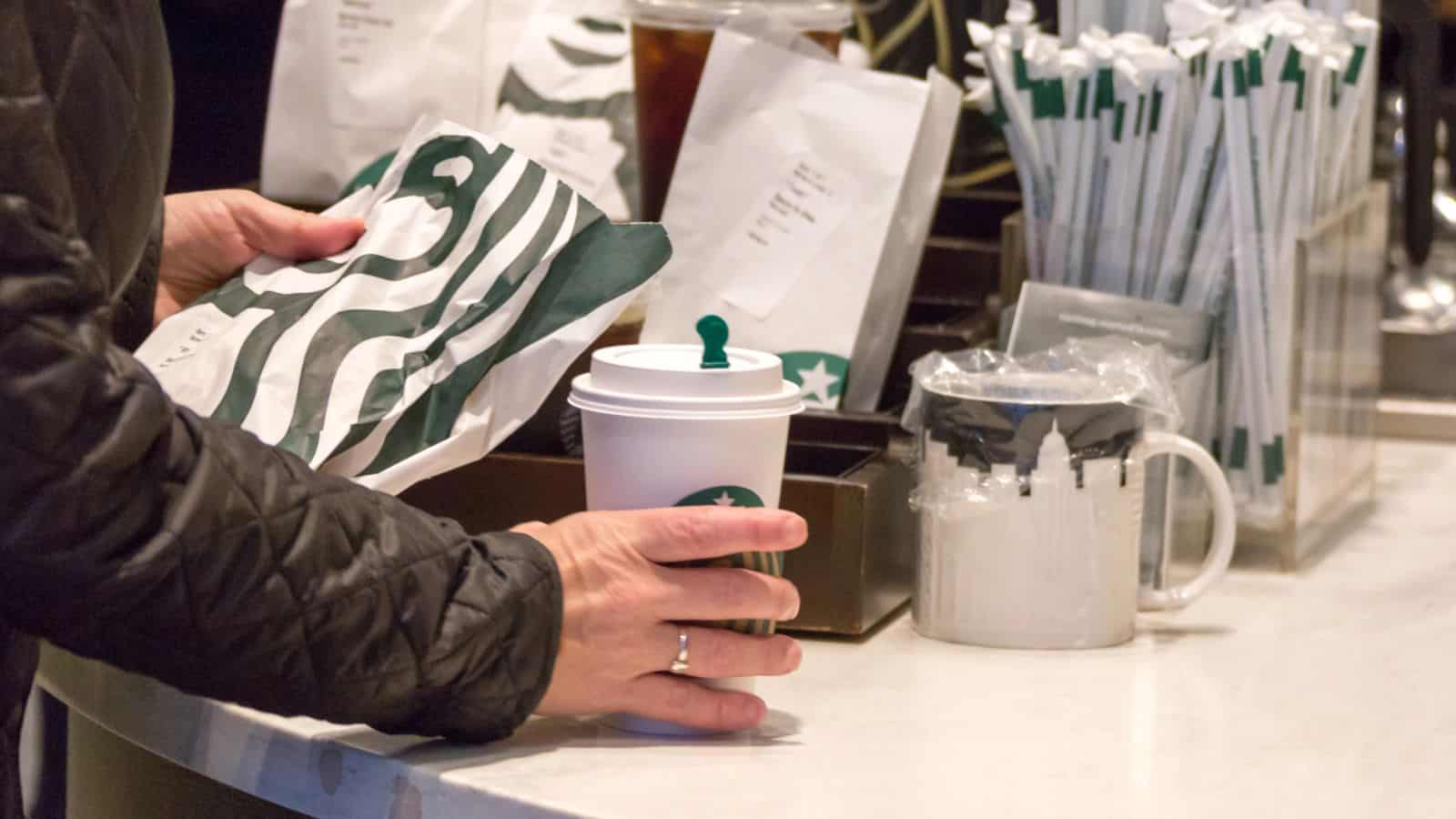 A person in a black jacket holds a Starbucks paper bag and a Starbucks cup at a counter. The counter also has a container of straws, a ceramic mug, and a plastic-wrapped item.