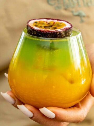 A person holds a layered yellow and green drink topped with a halved passion fruit, showcasing one of the essential fruits for home bartenders.